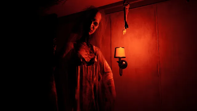 An old woman in a nightgown stands in a red-lit room