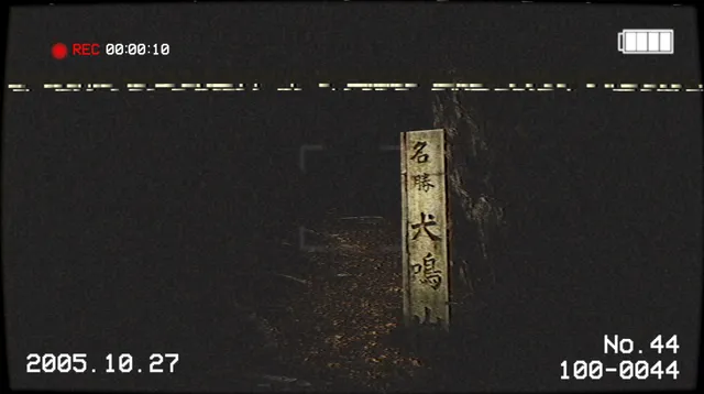 A wooden post marked Inunaki in Japanese beside a dark forest path, in a VHS camcorder frame