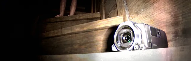 Jisatsu key art: a camcorder resting on old wooden stairs, a pair of bare legs standing in the dark at the top