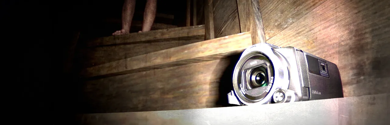 Jisatsu key art: a camcorder resting on old wooden stairs, a pair of bare legs standing in the dark at the top