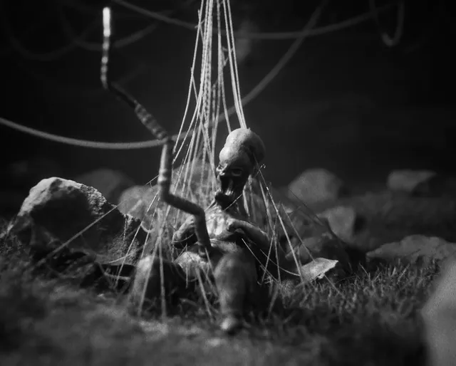 A skeletal stop-motion figure caught in hanging strings among rocks and grass