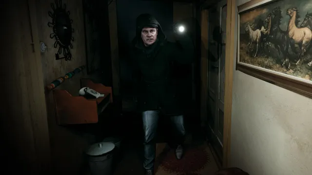 A man in a dark hooded coat with a flashlight in a narrow, cluttered hallway