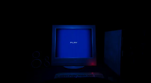 Play With Him screenshot: a blue CRT monitor showing the word PLAY, with speakers, a keyboard and a computer case in the dark