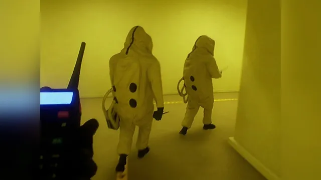Two figures in yellow hazmat suits walking down a yellow hallway, a walkie-talkie in the foreground