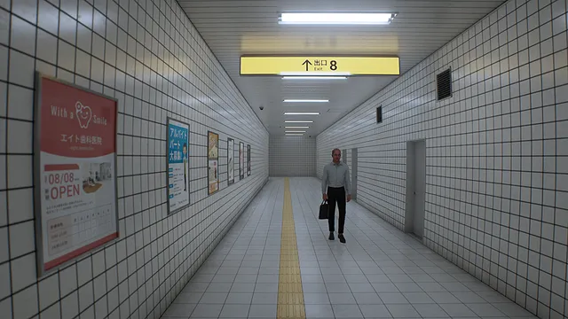 The Exit 8 screenshot: a man in a white shirt with a briefcase walks toward you down the tiled passage lined with posters