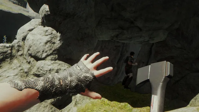 The Forest screenshot: first-person view of a hand in a chainmail glove and an axe near a cave mouth where a person stands