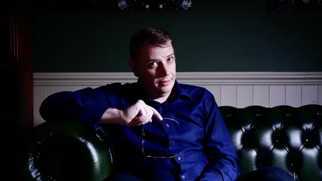 A man in a blue shirt on the couch, holding his glasses