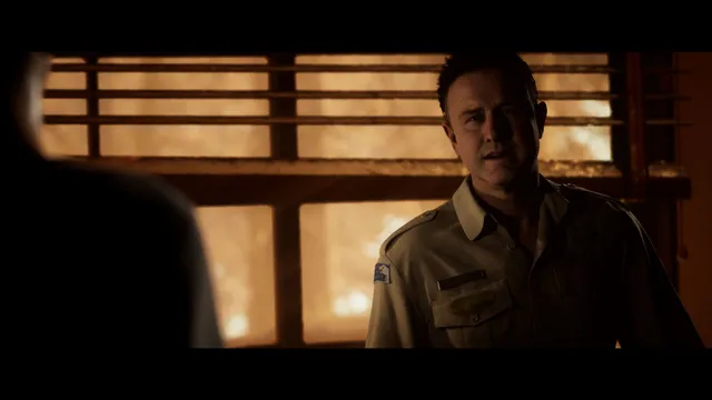 The Quarry screenshot: the camp's owner in a uniform shirt stands before a window glowing with fire