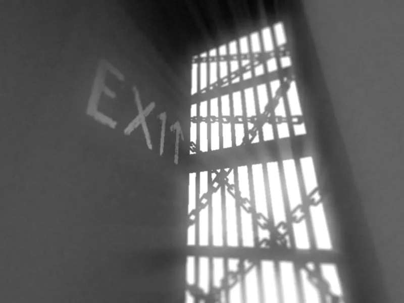 Which screenshot: in grainy greyscale, a barred window wrapped in chains lets in bright light, with the word EXIT and an arrow scrawled on the wall beside it